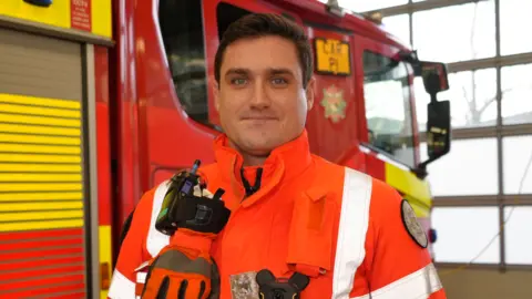 Shaun Whitmore/BBC Cameron Dowie who is standing next to a red and yellow marked fluorescent fire engine. He is looking directly at the camera and smiling and is wearing a fluorescent orange jacket. 