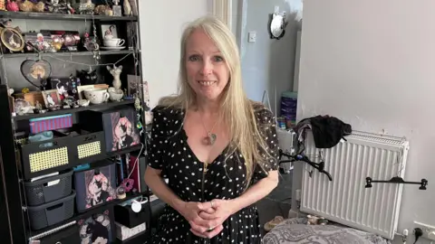 Maxine has long blonde hair and is wearing a black and white polka dot dress, with a silver necklace. She is standing in her living room, with shelves full of trinkets in the background.