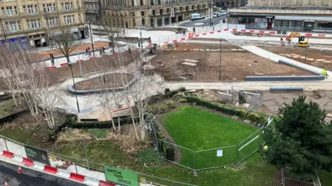 A view of an urban area being redeveloped into a park with green spaces and surrounded by red and whit barriers 