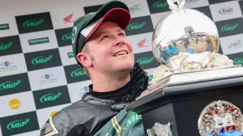 Daniel Ingham holding the Senior trophy while on the podium at the Manx Grand Prix. He is wearing black leathers and has two baseball caps on.