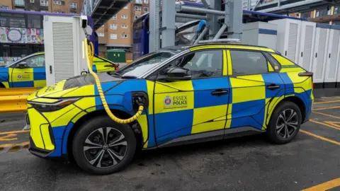 An Essex Police car charges up at an electric vehicle charging station at the First Bus depot in Basildon. The car is covered in blue and yellow squares, and there is a yellow and black charging cable reaching from a terminal across the bonnet of the vehicle and into its left-hand side.