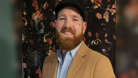 Anth Matthews Anth, a man with a long, brown beard and tattoos on his face. He is wearing a black flat cap, a brown jacket, and a blue button-up shirt. 