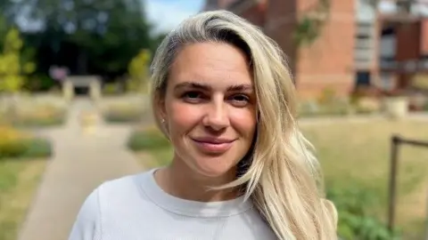 Jenna Robinson is smiling into the camera. She has long blonde hair and is wearing a white top. She is standing in a public garden which has wild flowers in it behind a council building in Beverley. 