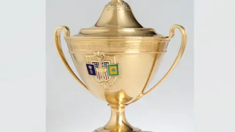 Gold trophy with dome lid and handles. A crest has badges on it 