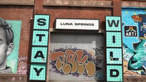 A grey metal shutter with orange graffiti is pulled over the entrance to Luna Springs. Murals are painted on the red brick wall around its entrance. Signs either side of the entrance say 'Stay Wild' in blue, with a white 'Luna Springs' sign above.