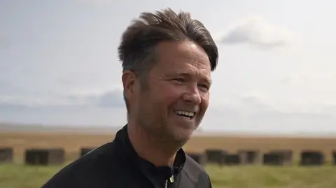 Conleth Maenpaa. He has short thick brown hair, slight stubble and is wearing a black jacket. Behind are green fields.