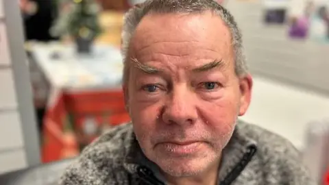 John is pictured in a community centre wearing a grey jumper. He has short greying hair, black eyebrows and grey stubble. 