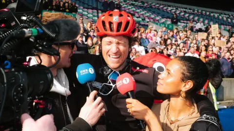Greg James, wearing a red cycle helmet and grimacing with exhaustion, puts his arms around colleagues Jack Saunders and Vic Hope, each of whom is holding a Radio 1-branded microphone. A cameraman trains his lens on Greg and a crowd can be seen looking on from the stands behind.