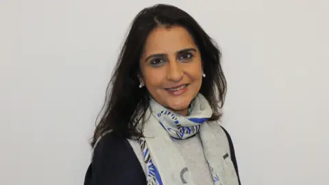 Dr Jyotsna Vohra Dr Jyotsna Vohra has long dark hair and is wearing a black top with a grey and blue scarf, she is stood slightly to the side so you can see her profile and is smiling at the camera.
