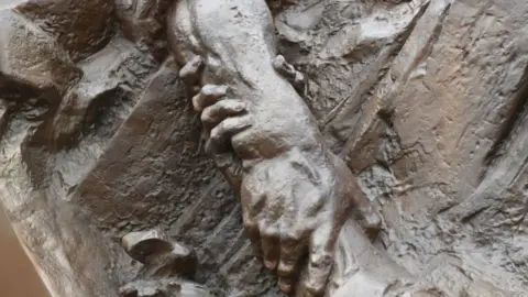 Ken Salmon Two arms in brass relief clutching each other as part of the memorial depicting a fisherman reaching out to rescue a PoW from the sea.