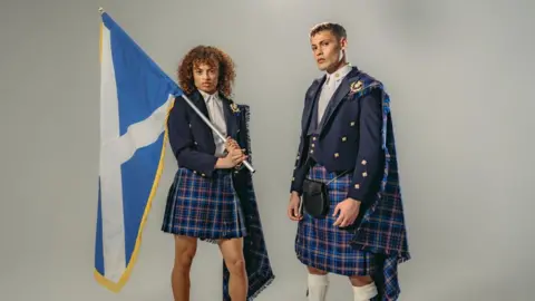 A male and female model in the Team Scotland uniform - blue tartan kilts and blue jackets. The female model is holding a saltire over her shoulders