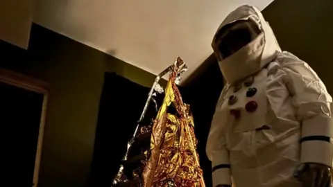 Deby McKnight A person wearing a white spacesuit and space helmet. There is a ladder behind them, which is wrapped in gold-coloured material. 