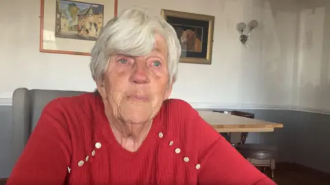 Blockley seated indoors at a table, facing the camera directly. She has short, neatly cut white hair and light-colored eyes, with natural facial lines visible around the eyes, cheeks, and mouth. She is wearing a red knit sweater that features decorative metallic buttons along the shoulder seams, and the forearms are resting on a surface just out of frame, suggesting a seated, slightly leaning-forward posture.