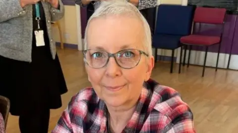 Sharon Hever is wearing a red, white and black top. She has short white hair and is wearing glasses. Some chairs and people, whose faces cannot be seen, are in the background.