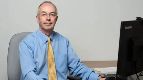 A picture of a man called Martin Tett who is the leader of Buckinghamshire Council. He is wearing a light blue shirt with a yellow tie and black dots and is sat at a desk with a computer monitor on his left.