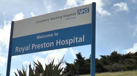 A blue and white sign reading 'Welcome to Royal Preston Hospital' in front of green bushes and and blue cloudy sky
