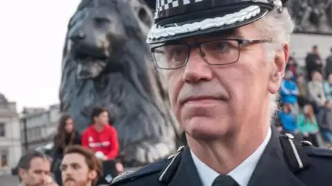 Alamy Julian Bennett wearing glasses and a police uniform in Trafalgar Square with crowds of people behind him