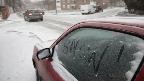 In the foreground, a parked red car sits at the side of a street with "Snow" scraped in the snow on the passenger window. Further away, a grey car drives on a snowy road surface with its brake lights on.