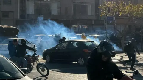 EPA Một đám khói hơi cay bao phủ con đường khi ô tô và xe máy đi qua, ở Tehran (25/12/2025)