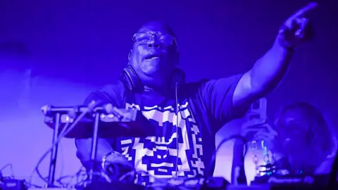 Getty Images Carl Cox behind the decks at a shiw. The stage is bathed in purple lighting and he has an arm in the arm pointing at the crowd. 