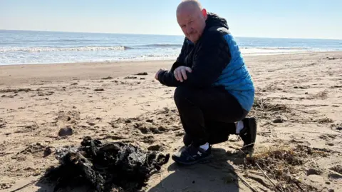 Kevin Shoesmith/BBC Steven Davison, wearing a blue and navy coat and joggers, on the golden sands of a beach with a blue sea and sky behind him. He is looking at a large black mound comprising molten pellets used to make plastic. 