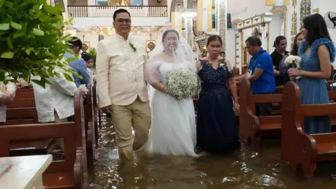 A igreja de Barasoain percorre a igreja inundada. Ela usa branco e segura um grande buquê de respiração do bebê