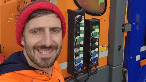 A smiling man with brown hair, beard and a red woolly hat takes a selfie in front of an orange bin truck. He wears an orange hooded top. Dials in green, black, light blue and dark blue run vertically in two channels, attached to what looks like telephone cables.