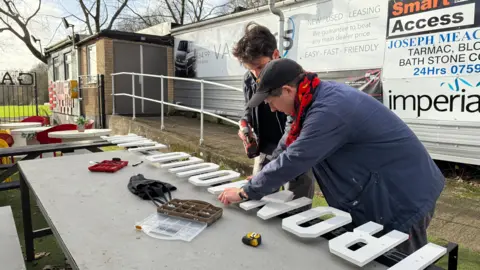 Mr Borg and Mr Kelham are wearing the same outfits as in the previous photo; however, they are off the pitch. They are standing next to a table with a large sign that reads 'Ironmould Lane' in white letters and are drilling the letters into a board.