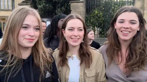 Three young women are smiling at the camera on a busy street in Oxford. They all have long hair and are standing in front of a crowd.
