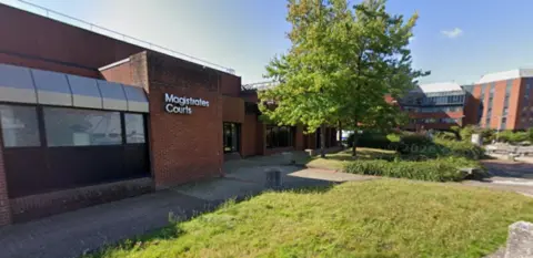 The exterior of a low-rise brick building. The words 'Magistrates’ Courts' are displayed in white lettering ear the main entrance.
The building is constructed primarily of red-brown brick and has a flat roof. Large windows line the front of the structure. The main entrance is set slightly back from the front edge of the building and appears to be accessed from a paved walkway.
