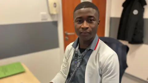 BBC Benjamin is sitting at a desk, looking towards the camera. He is wearing a blue top, a white jacket and an NHS lanyard. The background is a blurred medical room.