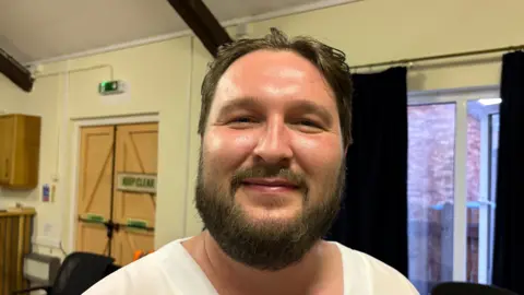 Martin Heath/BBC Matt Hensey with a beard wearing a light-coloured top stands inside a community hall. Behind the person are dark curtains, chairs, wooden double doors with a "KEEP CLEAR" sign, and exposed ceiling beams