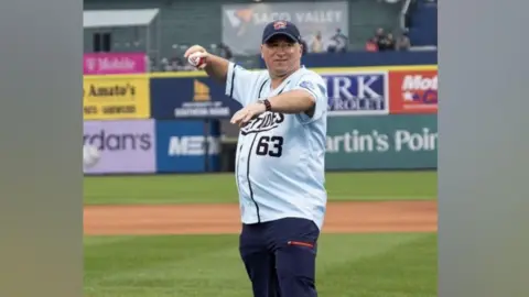 John Lewis threw the ceremonial first pitch in front of thousands of fans. He is wearing a pale blue a baseball top with dark letter on it and dark coloured shorts and a dark baseball cap. He is standing on the pitch with sports stands behind him.