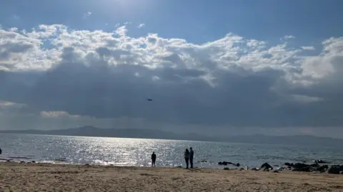 Three people standing on Buncrana beach in County Donegal are looking out to sea on a sunny day and in the sky a coast guard helicopter can be seen searching the water for swimmers who have been reported missing