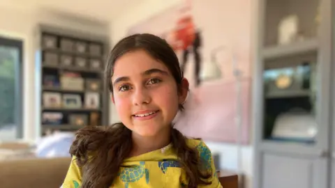 An eleven year old girl with long brown hair smiles at the camera with the background room blurred.