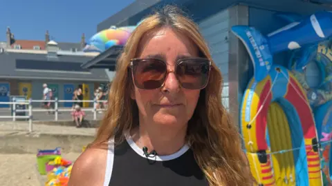 Debbie Rait is standing next to her stall on the beach. She has brown shoulder length hair and is wearing large sunglasses. She is wearing a black vest.
