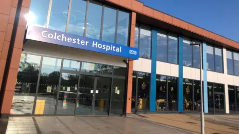 ESNEFT The entrance to Colchester Hospital, which is a two-storey building with a glass facade. It has a blue banner that says Colchester Hospital above the entrance doors.