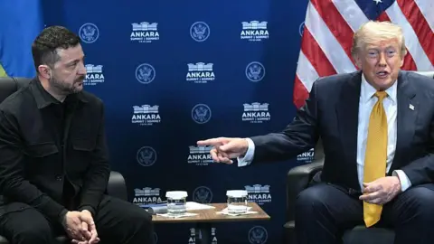 Zelensky and Trump seated at a news conference at the Nato summit in Ankara, Turkey. Trump is speaking to the cameras and pointing at Zelensky, who is looking towards him.