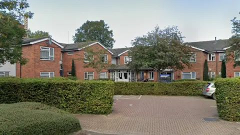 A small carpark is at the front of the picture, that has rows or green bushes around it. In the background there are lots of rows of buildings, with multiple windows across two stories. 