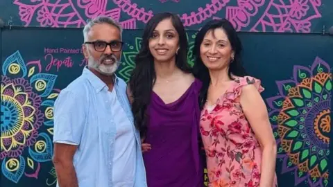 Supplied The picture shows a male and two females stood in front of a multicoloured mandala background. The male is wearing light blue, while one female is wearing dark purple and the other a floral pink dress. 