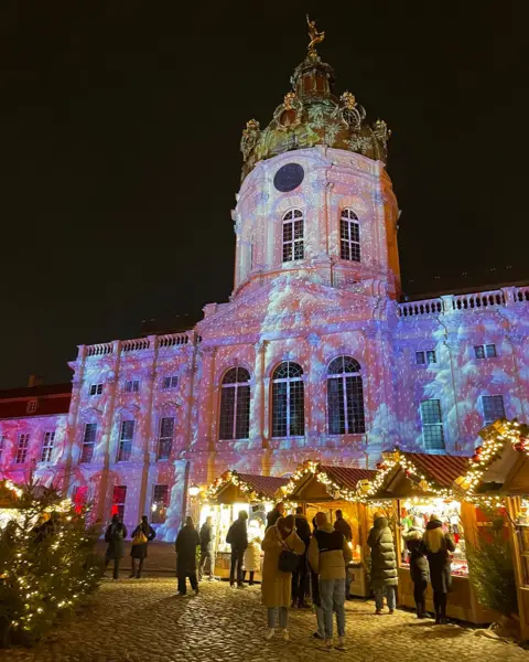 A pink projection onto a historical palace building in Berlin, Germany, with wooden huts lined with fairy lights and Christmas trees in the foreground in front of the palace, as people mingle at Christmas market stalls.