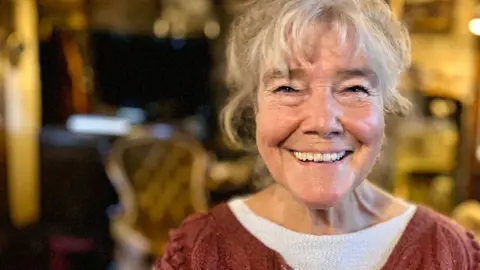 Christine Adams looks direct at the camera and smiles broadly. She has white hair and is in the sitting room of a house. She is wearing a plum-coloured cardigan over a white sweater. 