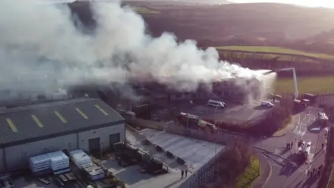 Firefighters on an aerial ladder platform try to douse the flames of a large fire at a school in Devon with water. Plumes of smoke are rising into the sky.
