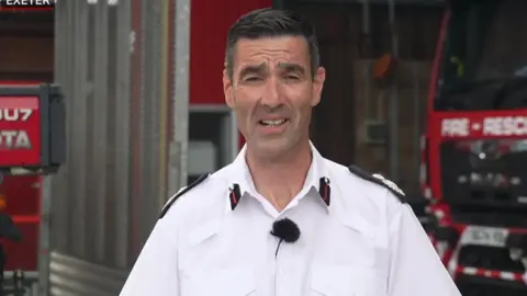 The Devon and Somerset fire's services's assistant chief is wearing a white shirt with black epaulettes. He is standing in front of a red fire engine.