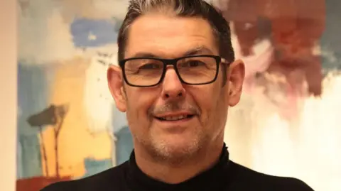 Handout Darren Diprose wearing a black turtle neck jumper and glasses stands smiling. There is a colourful painting in the background.