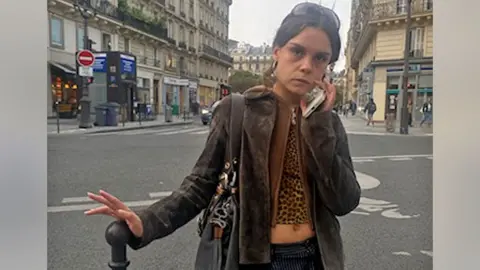 Paola Goddard brown hair young woman stands in the street with her hands on top of the bollard. She wears a brown jacket and a leopard print top. She has her phone on her face and has a serious expression