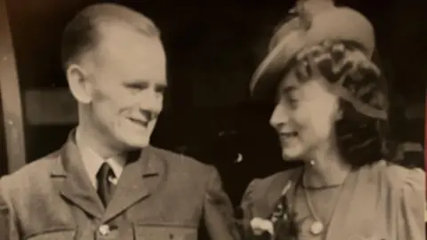 Andrea Morrison A sepia-toned photograph shows two individuals standing side by side, dressed in formal attire from the mid-20th century. The person on the left is wearing a military-style uniform with a buttoned jacket and tie. The person on the right is wearing a light-colored dress with puffed sleeves and a decorative brooch, along with a wide-brimmed hat adorned with flowers. A corsage of flowers is pinned to the dress. The image has a classic, aged appearance.