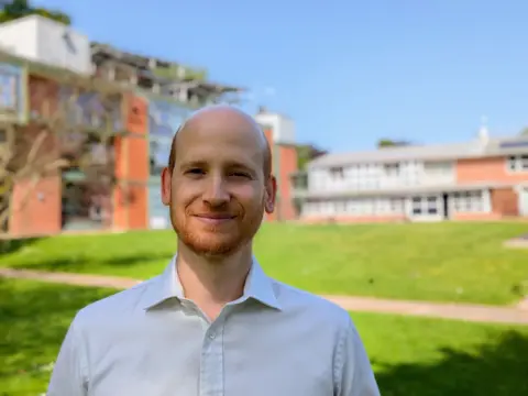 Supplied A man with a ginger beard wearing a white collared shirt, smiling and stood in front of a grassy courtyard.