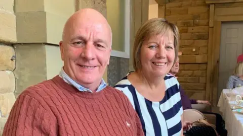 A middle-aged couple posing for a picture during dinner with a larger group of people. The man, on the left, is wearing an orange jumper with a blue shirt underneath. The woman, on the right, is wearing a navy and white striped top