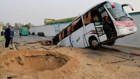 Two men look at a large bus which has tipped into a crater, burying the back half of the vehicle